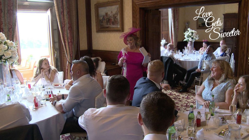 A still from their wedding video showing the wedding reception room with white and blush pink flowers during the mother of the bride speech at a wedding in armathwaite hall hotel in Keswick