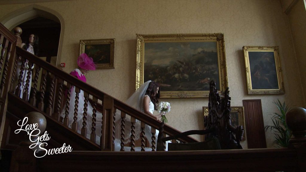 The wedding videographer videos the Bride and her Mum making their way down the old staircase at Armthawaite Hall hotel in Keswick for their wedding video in Cumbria. Traditional old paintings hang behind making this a very classic bridal shot